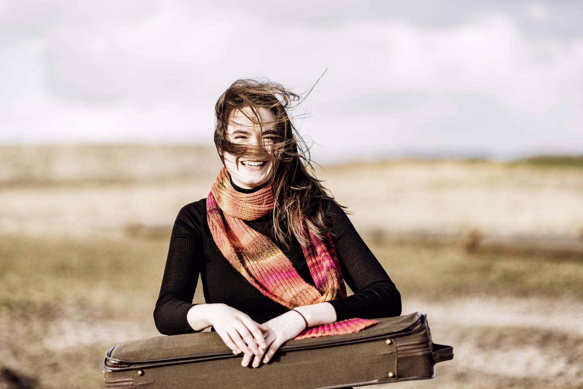 Hadewych outside with colorful scarf holding violin case, with hair across her face
